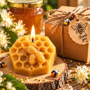 Op de afbeelding: Een goudgele bijenwaskaars in de vorm van een honingraat met een bijenreliëf aan de bovenkant en een brandende lont. De kaars staat op een houten schijf naast een bruine geschenkdoos met touw en een label met een bijenillustratie en de tekst thank you.