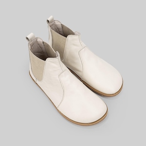 May include: A pair of cream-coloured Chelsea boots. The boots feature a smooth leather upper, elastic side panels, and a pull tab at the back. The soles are light brown. These boots are suitable for everyday wear.