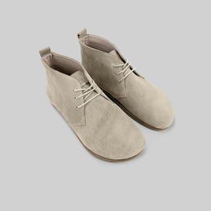 Puede incluir: Un par de botines chukka de ante beige claro con cordones blancos. Los botines tienen una puntera redondeada y un perfil bajo, adecuados para ropa informal. Las suelas son de color marrón en contraste.
