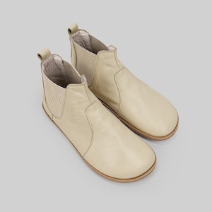 May include: A pair of cream-coloured Chelsea boots. The boots feature a rounded toe, elastic side panels, and a pull tab at the back. The soles are light brown. The boots are made of a smooth leather-like material.