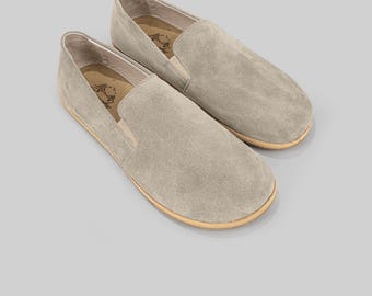 May include: A pair of light gray suede slip-on shoes. The shoes have a rounded toe and a tan sole. The interior of the shoe has a printed design. The shoes are displayed on a light gray background.
