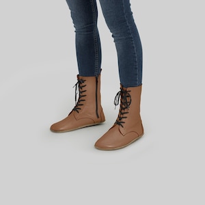 Puede incluir: Botas de cuero color canela con cordones y cremalleras negras. Las botas tienen suela plana y altura media de la pantorrilla. Las botas se llevan con vaqueros azules.