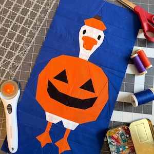 May include: A rectangular fabric quilt block featuring a pieced design of a white goose wearing an orange pumpkin costume. The goose has an orange beak and feet, while the pumpkin body has black triangular eyes and a wide black mouth.