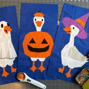 Può includere: Tre blocchi trapuntati rettangolari in tessuto raffiguranti oche bianche su sfondo blu. Un'oca indossa un costume da fantasma con lenzuolo bianco, una indossa un costume da zucca arancione con faccia da jack-o'-lantern e una indossa un cappello da strega viola.