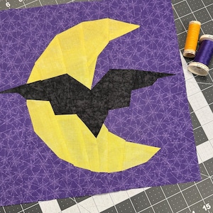 May include: A Halloween-themed fabric square with a yellow crescent moon and a black bat on a purple spiderweb-patterned background. Two spools of thread in orange and purple are visible.