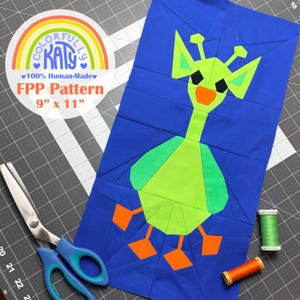 May include: A fabric quilt block featuring a stylised green giraffe on a solid blue background, with orange feet and beak, and black eyes. A circular logo reads Colorfully Katy 100% Human-Made FPP Pattern 9" x 11". Includes blue scissors and thread.