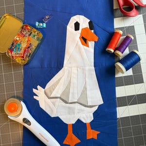 May include: A fabric quilt block featuring a white and grey seagull with an orange beak and feet on a solid blue background. The block is surrounded by sewing tools including red scissors, a rotary cutter, three spools of thread, and a metal tin.