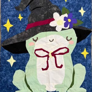 May include: A whimsical quilt featuring a frog wearing a witch hat adorned with flowers. The frog is light green with a white belly and a burgundy bow. The background is a dark blue with yellow star accents. Text reads "Pattern Tester: Joah Bingert."