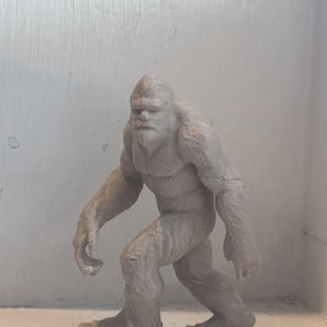 May include: A light brown figurine of a Bigfoot creature, sculpted in a walking pose. The figure has a detailed face with a beard and fur-like texture on the body. The figurine is standing on a textured base.