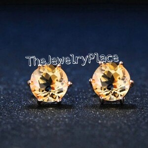 May include: A pair of round, golden-yellow gemstone stud earrings. The earrings are set in a rose-gold colored metal and are displayed against a dark blue background. The text "TheJewelryPlace" is visible.