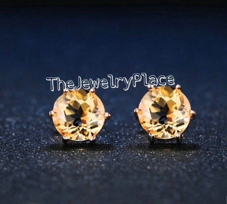 May include: A pair of round, golden-yellow gemstone stud earrings. The earrings are set in a rose-gold colored metal and are displayed against a dark blue background. The text "TheJewelryPlace" is visible.
