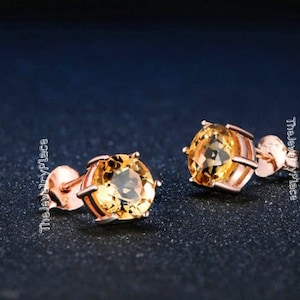 May include: A pair of stud earrings featuring round, golden-yellow gemstones set in rose gold metal. The earrings have a classic, elegant design, perfect for everyday wear or special occasions. The Jewelry Place is printed on the side.