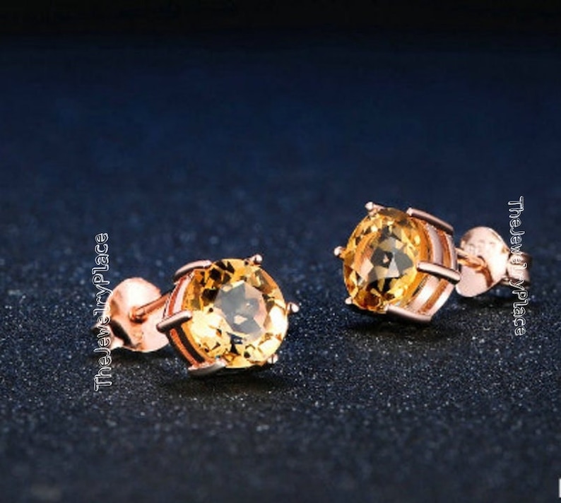 May include: A pair of stud earrings featuring round, golden-yellow gemstones set in rose gold metal. The earrings have a classic, elegant design, perfect for everyday wear or special occasions. The Jewelry Place is printed on the side.