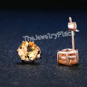 May include: Rose gold stud earrings with round, faceted, yellow gemstones. The earrings are set in a four-prong setting and have a post and butterfly backing. The text "TheJewelryPlace" is visible in the image.