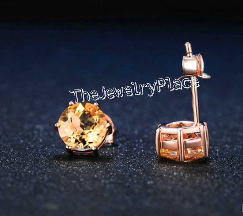 May include: Rose gold stud earrings with round, faceted, yellow gemstones. The earrings are set in a four-prong setting and have a post and butterfly backing. The text "TheJewelryPlace" is visible in the image.