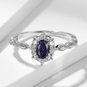 Puede incluir: Un anillo de plata con una piedra preciosa azul engastada en un delicado diseño floral. La banda es retorcida y lleva la inscripción "The Jewelry Place" y "S925".