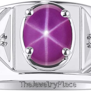 Könnte beinhalten: Ein silberner Ring mit einem großen ovalen, violetten Stern-Saphir-Edelstein und zwei kleineren Diamant-Akzenten. Der Ring hat eine strukturierte Band und die Worte "The Jewelry Place" sind auf der Innenseite der Band eingraviert.