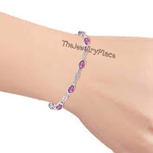 May include: A silver bracelet with pink oval-shaped gemstones and small clear round gemstones. The bracelet is on a person's wrist. TheJewelryPlace is written on the wrist.