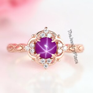 Può includere: Anello in oro rosa con una gemma di rubino a stella. L'anello presenta un design floreale attorno alla pietra centrale, impreziosito da piccole gemme chiare. La fascia ha un motivo dettagliato e strutturato. Gioielli di TheJewelryPlace.