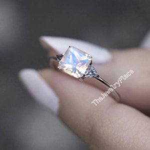 Può includere: Un anello d'argento con una grande pietra di luna di forma quadrata e due pietre triangolari più piccole su entrambi i lati. L'anello è su un dito.