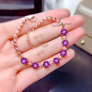 May include: A silver bracelet with a rose gold finish features a row of purple star-shaped gemstones set in flower-shaped settings. The bracelet has a lobster clasp closure.