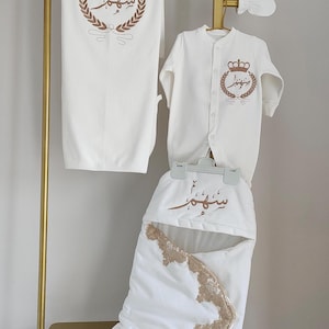 Puede incluir: Un conjunto de ropa de bebé blanco con bordados dorados. El conjunto incluye un saco de dormir, un body, un gorro y manoplas. Cada artículo presenta un diseño de corona y corona de laurel con escritura árabe. Los artículos se exhiben en un estante dorado.