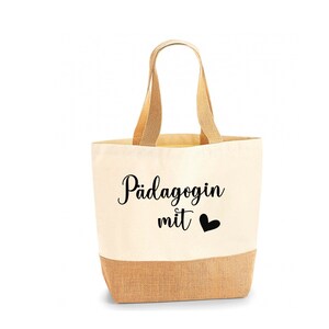May include: A beige tote bag with a natural jute bottom and handles. The bag features the text "P&auml;dagogin mit" and a black heart symbol, likely a gift item.