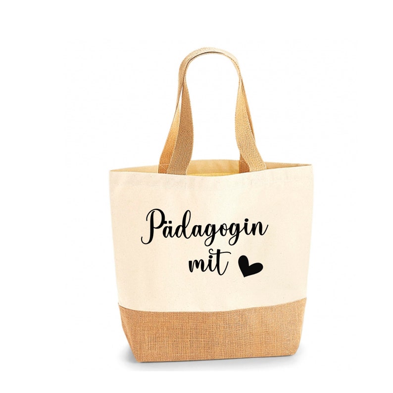 May include: A beige tote bag with a natural jute bottom and handles. The bag features the text "P&auml;dagogin mit" and a black heart symbol, likely a gift item.