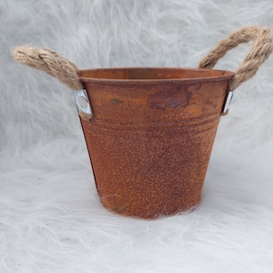 Könnte beinhalten: Ein kleiner, rostiger Metalleimer mit zwei Seilgriffen. Der Eimer hat ein verwittertes, strukturiertes Aussehen mit sichtbaren Rostspuren. Die Seilgriffe sind mit Metallbügeln befestigt. Dieser dekorative Eimer eignet sich für kleine Pflanzen oder als rustikales Wohnaccessoire.