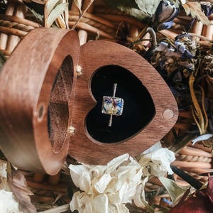 Op de afbeelding: Een hartvormige houten ringdoos, open om een zilveren ring met een vierkante, kleurrijke edelsteen te onthullen. De doos staat tegen een achtergrond van gedroogde bloemen en geweven materiaal, wat een rustieke en romantische uitstraling creëert.