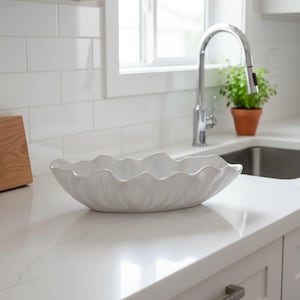 Può includere: Una ciotola ovale in ceramica bianca con bordo smerlato e ondulato e finitura lucida. La ciotola presenta un design testurizzato a forma di petalo che si irradia dal centro verso i bordi, appoggiata su una superficie liscia del piano cucina.