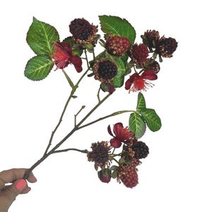 May include: A decorative artificial branch featuring dark purple and red blackberries, green serrated leaves, and small red flowers with thin, spiky petals. The branch has a brown stem and is arranged with multiple clusters of berries and foliage.
