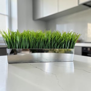 Puede incluir: Una jardinera rectangular con un acabado metálico plateado pulido y reflectante que contiene una disposición densa de briznas de hierba artificial de color verde vibrante, creando una apariencia uniforme y texturizada a lo largo del recipiente.