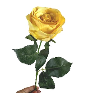 Könnte beinhalten: Eine einzelne künstliche Rose mit leuchtend gelben Blütenblättern, die in einer vollen, geschichteten Blüte angeordnet sind. Die Blume ist an einem schlanken grünen Stiel befestigt, der mehrere dunkelgrüne, gezackte Blätter mit sichtbaren Blattadern aufweist.