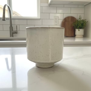 May include: A cylindrical ceramic planter with a speckled off-white glaze finish. The vessel features a smooth, straight-sided body that tapers slightly toward a circular base, creating a clean, uniform profile for holding small indoor plants.