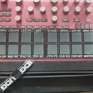Puede incluir: Un dispositivo rosa de producción de música electrónica con numerosos botones, perillas y pads. El dispositivo tiene un panel negro con etiquetas de texto como "SWING", "LENGTH" y "KEY". Una pegatina negra de UDG es visible.