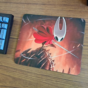 May include: A square mousepad featuring a character from the video game Hollow Knight. The character, Hornet, is depicted with a red cape, a white mask, and a needle. The background is a fiery, orange and red landscape.