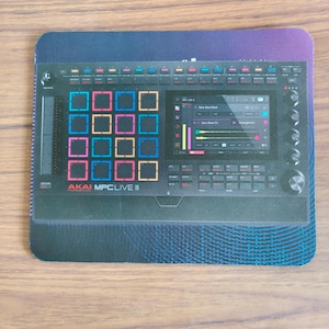 May include: A rectangular mousepad featuring a graphic print of an Akai MPC Live III music production controller. The black device displays a grid of colorful square pads, a digital touchscreen interface, various control knobs, and buttons.