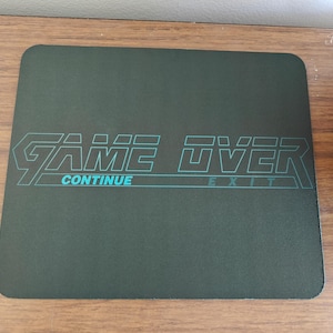 Puede incluir: Una alfombrilla de ratón cuadrada de color verde oscuro con las palabras "GAME OVER" en una fuente retro, con "CONTINUE" y "EXIT" debajo en color turquesa. La alfombrilla está sobre una superficie de madera.