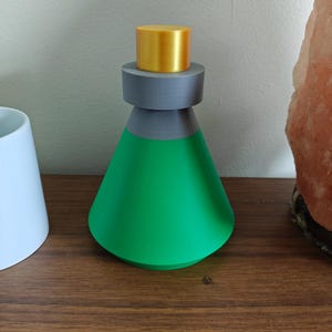 May include: A green, cone-shaped bottle with a gray neck and a gold cap. The bottle is sitting on a brown wooden surface. The bottle is next to a white cylindrical object.