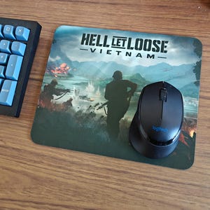 May include: A rectangular mousepad featuring the game title "HELL LET LOOSE - VIETNAM" with a war scene graphic. A black Logitech mouse sits on the pad next to a blue-keyed keyboard.