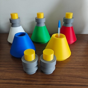 Puede incluir: Una colección de recipientes cónicos coloridos con tapas amarillas y bases grises. Los recipientes son de varios colores, incluyendo blanco, verde, azul, rojo y amarillo. Un recipiente amarillo contiene un bolígrafo azul. Están dispuestos sobre una superficie de madera.