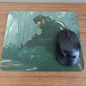 May include: A black Logitech wireless mouse sits on a rectangular mouse mat. The mat displays a green and grey illustration of a man with a rifle in a jungle. The mouse mat is on a wooden surface.