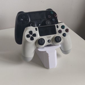 May include: Two PlayStation 4 controllers, one black and one white, are displayed on a white stand. The controllers are positioned side-by-side, with the black controller on top of the white controller.