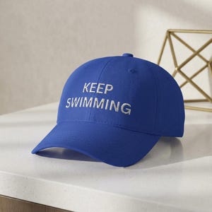 May include: A royal blue baseball cap with the embroidered text "KEEP SWIMMING" in white. The cap has a curved brim and a rounded crown. The hat is made of a soft fabric and is suitable for casual wear.