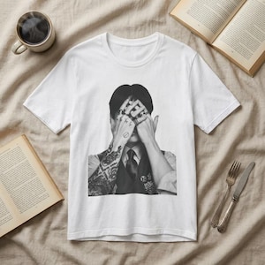 Op de afbeelding: Wit t-shirt met een zwart-wit afbeelding van een persoon met tatoeages en ringen die hun ogen bedekken. De persoon draagt een vest en stropdas. Een kopje koffie en open boeken staan op de achtergrond.