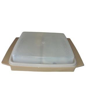 May include: A tan-colored, rectangular food storage container with a clear, square lid. The container has a slightly raised edge and a flat base. The lid appears to be made of a translucent plastic material.