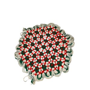 May include: A round, handmade trivet with a floral pattern. The trivet features a green background with white and red flower-like designs. The edges are scalloped and finished with a cream-colored yarn.