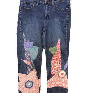 May include: Blue denim jeans with colorful patchwork designs. The patches include stars, triangles, and other shapes in various patterns and colors, including red, pink, green, and purple. The jeans are hanging on a black hanger.