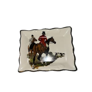 May include: A decorative, off-white ceramic dish with a wavy edge and a black and gold trim. The dish features a detailed illustration of two riders on horseback, with a hunting scene.